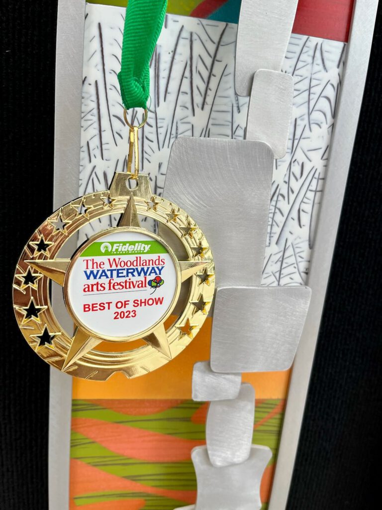 a beautiful medal, with gold star stylings, from The Woodlands Waterway Arts Festival, hanging on an original metal wall sculpture by Cherie Haney