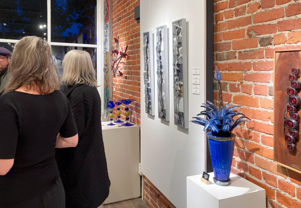 Three muted color wall sculptures hanging in a trio in between original glass sculptures in the Chris Nordin Studio Gallery.  Guests mingle nearby.