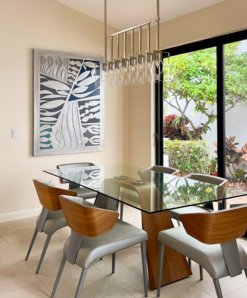 A large abstract metal art piece hangs on the wall behind a glass dining room table. Natural light comes from a door wall to the right. A modern chandelier with multiple lights, unlit, hangs over the table.