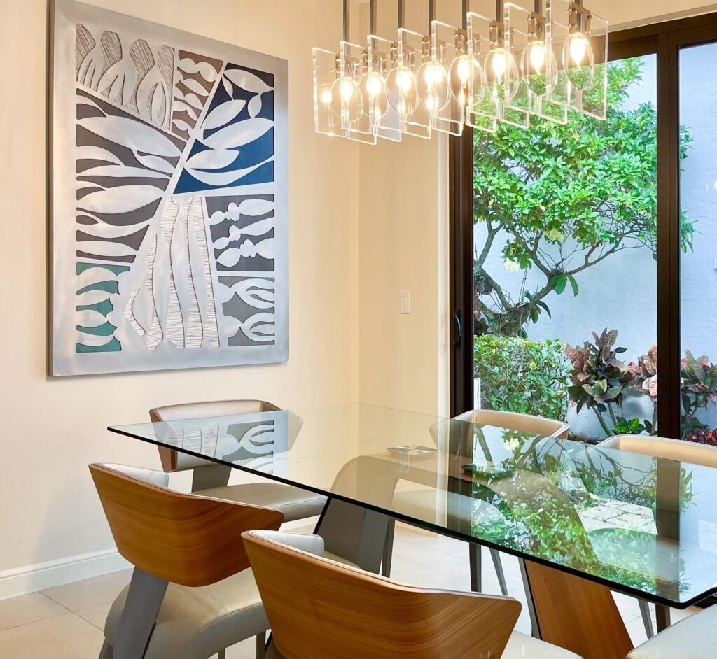 A large abstract metal art piece hangs on the wall behind a glass dining room table. Natural light comes from a door wall to the right. A modern chandelier with multiple lights hangs over the table.