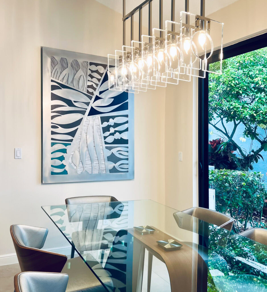 A large abstract metal art piece hangs on the wall behind a glass dining room table. Natural light comes from a door wall to the right. A modern chandelier with multiple lights hangs over the table.