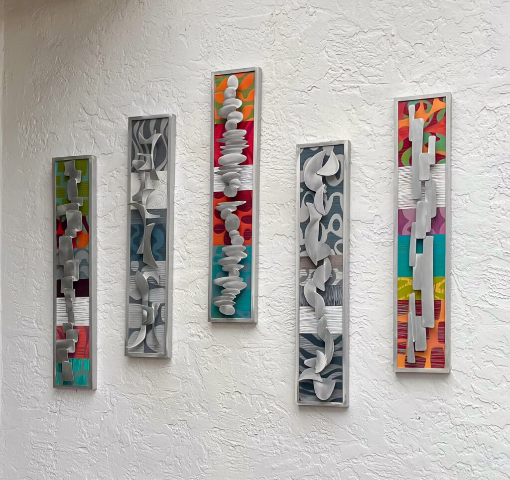 A grouping of five small narrow mixed media wall art sculptures hang on an exterior wall. The artwork is colorful and contains abstract aluminum shapes.