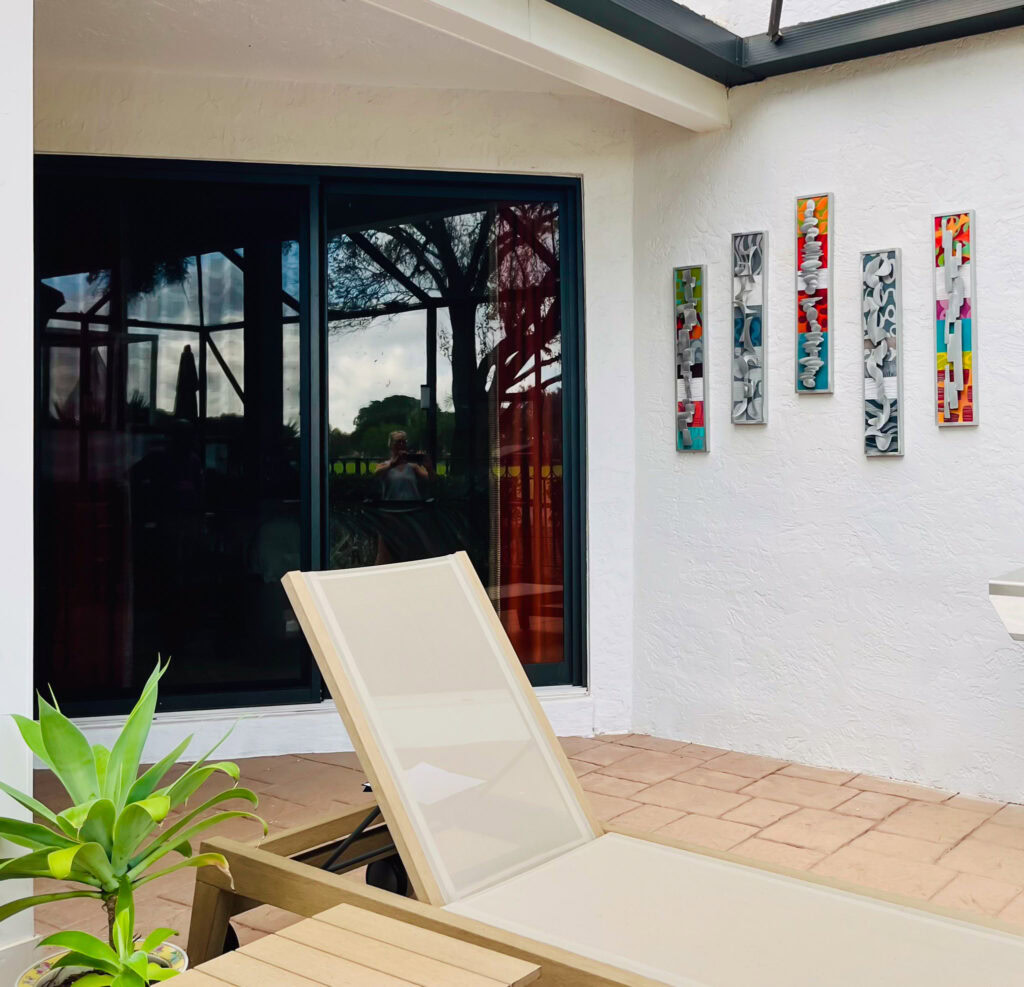 A grouping of five small narrow mixed media wall art sculptures hang on an exterior wall. A lounge chair sits in the foreground and a large door wall in the back left of the frame. The artwork is colorful and contains abstract aluminum shapes.