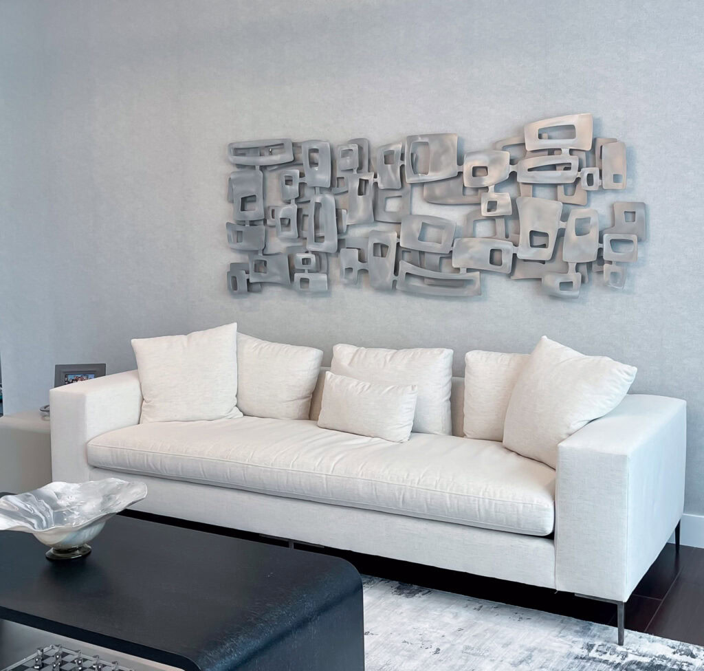 freeform square abstract metal wall art sculpture hanging over a white couch. A modern chandelier hangs from the ceiling, a black table is in the foreground, and natural light comes through a large window to the right. The sculpture is brushed aluminum pieces grouped and layered into an abstract dimensional field of square and rectangular shapes.