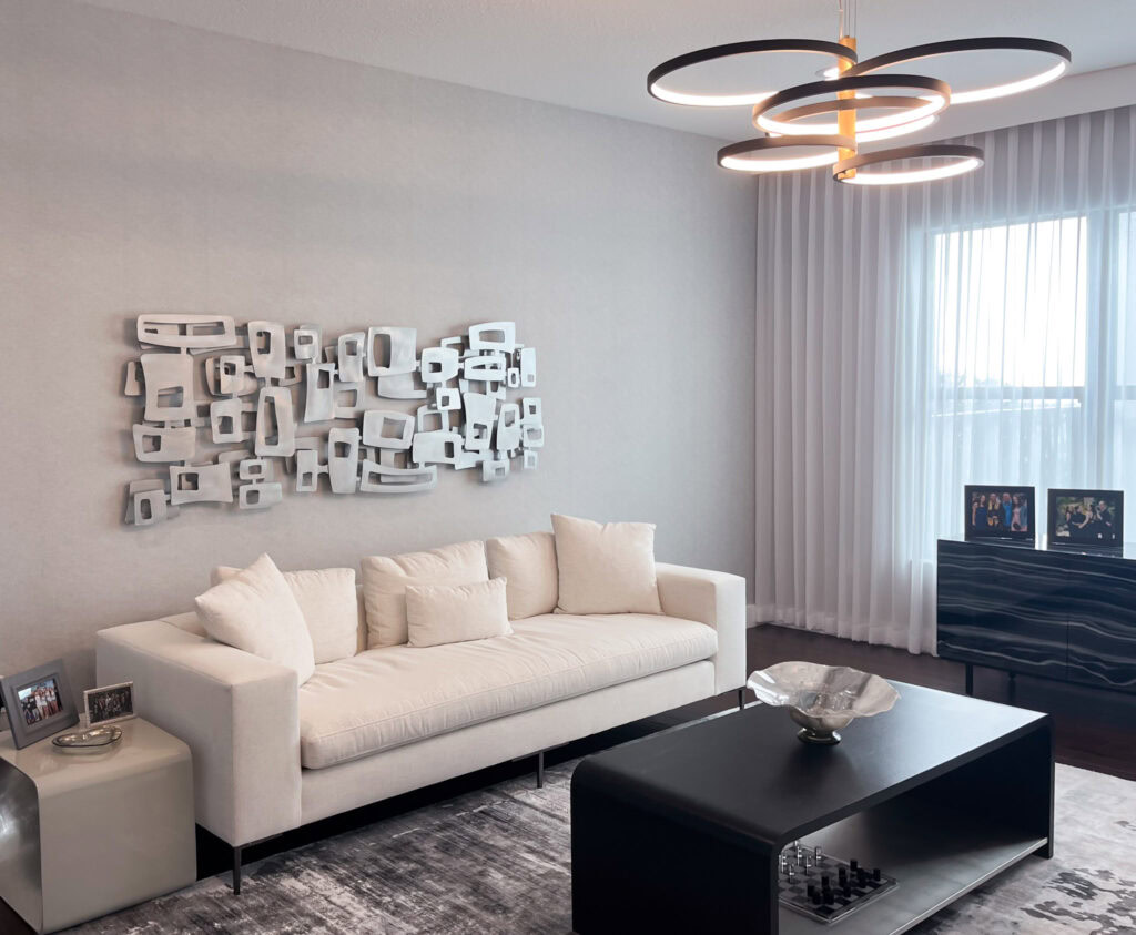freeform square abstract metal wall art sculpture hanging over a white couch. A modern chandelier hangs from the ceiling, a black table is in the foreground, and natural light comes through a large window to the right. The sculpture is brushed aluminum pieces grouped and layered into an abstract dimensional field of square and rectangular shapes.